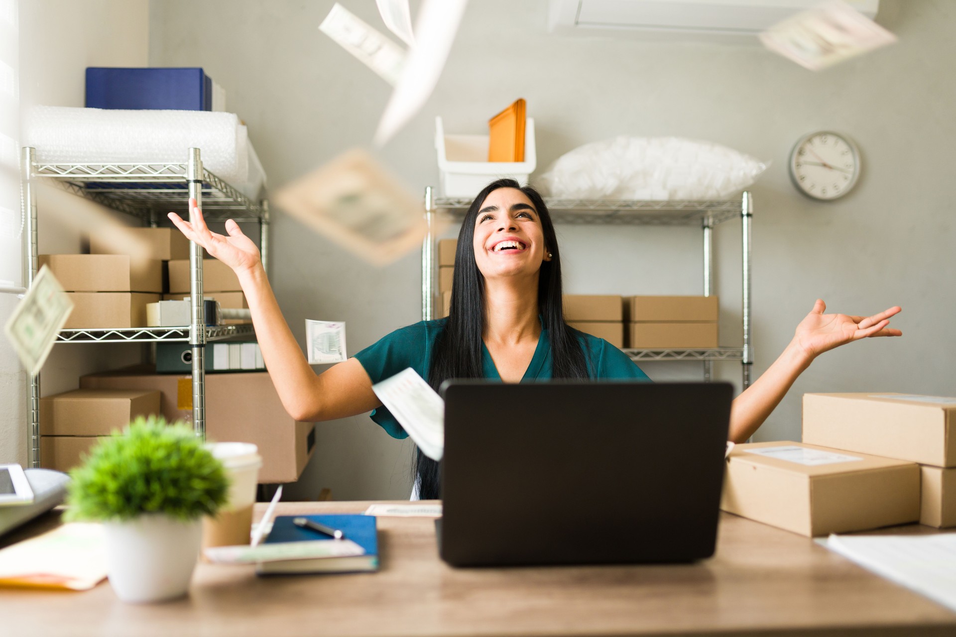 Excited businesswoman throwing money in the air while working on laptop in her warehouse, celebrating success of her small business